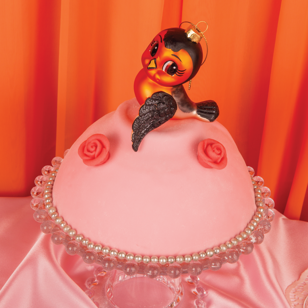 Dompap julekule fra HEIM Studio – håndmalt glassfigur i rød og svart med store øyne og vipper. Nostalgisk og sjarmerende julefugl. Plassert på en rosa marsipankake med oransje bakgrunn.