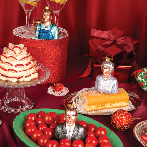 Julekuler i glass fra HEIM Studio inspirert av The Julekalender – Olaf Sand med pipe, Gjertrud Sand med perlekjede og Benny/Nåsåen i dress. Håndmalte figurer plassert i et julepyntet bordmiljø.